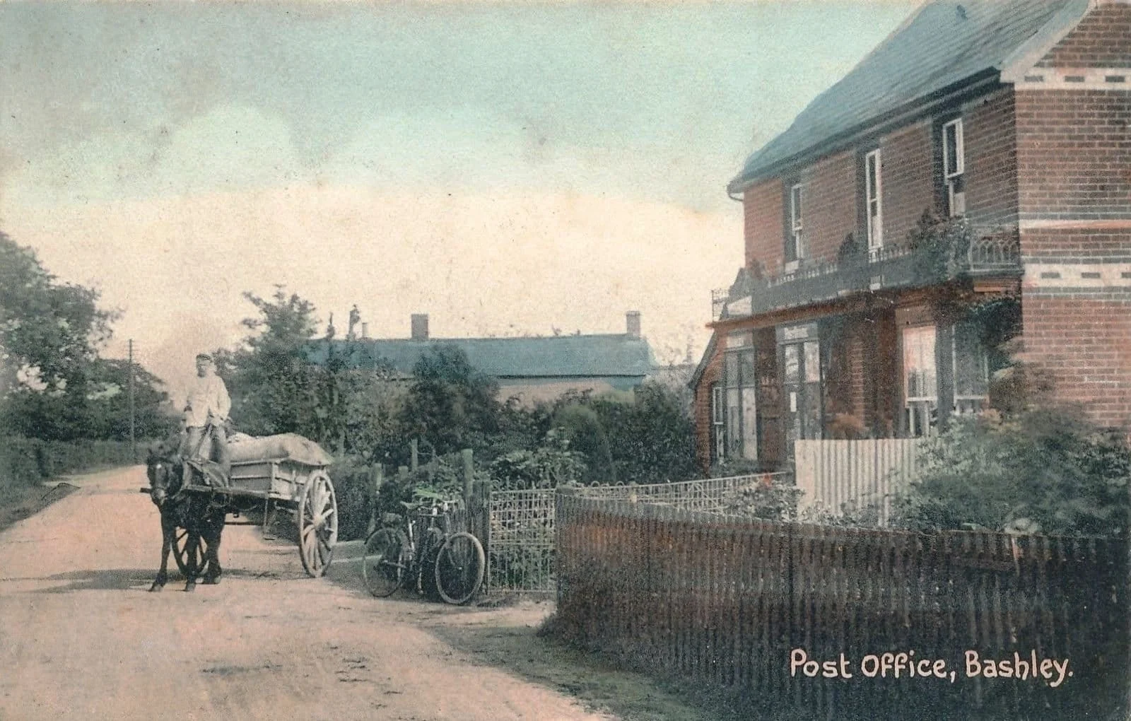 A vintage photo of the Post Office in Bashley shows a brick building with a fenced yard. A man sits on a horse-drawn cart on a dirt road, with two bicycles resting against the fence.