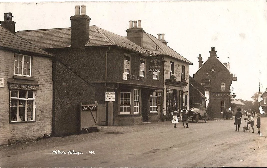 A vintage black-and-white photo of Milton Village shows early 20th-century buildings, a garage, shops, and people in period clothing walking or standing on a wide street, with a dog near several children.