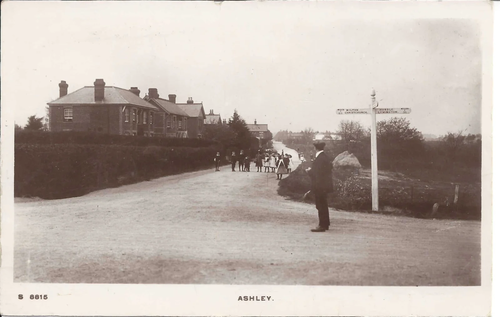 A vintage photo shows a quiet village road in Ashley, with houses on the left, a group of people gathered in the distance, and a man standing by a signpost at a crossroads. Trees and hedges line the scene.