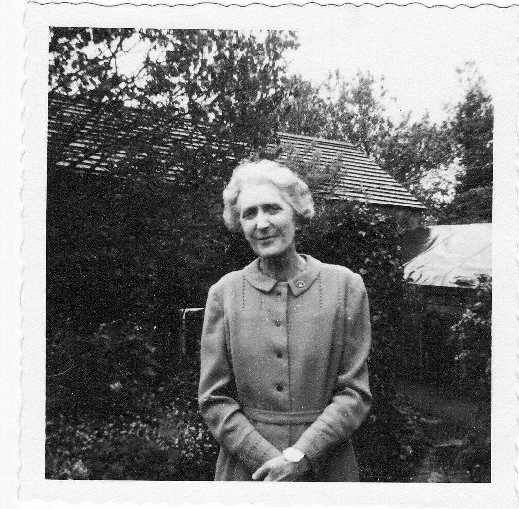 Black and white photo of an older woman with short curly hair, wearing a buttoned-up dress, standing outside in a garden with trees and a house in the background.