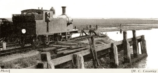 A vintage steam locomotive with a driver in the cab stands on a railway track beside a wooden quay, near a body of water in a rural landscape.