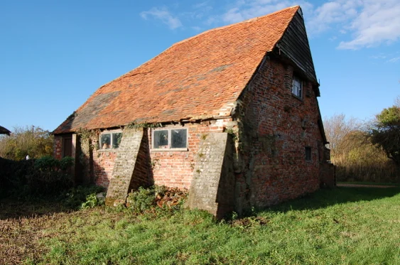 A weathered brick barn with a red tiled roof stands on grassy ground. The building has two large support buttresses and small, rectangular windows. Some bricks show signs of age and moss; the sky is clear and blue.