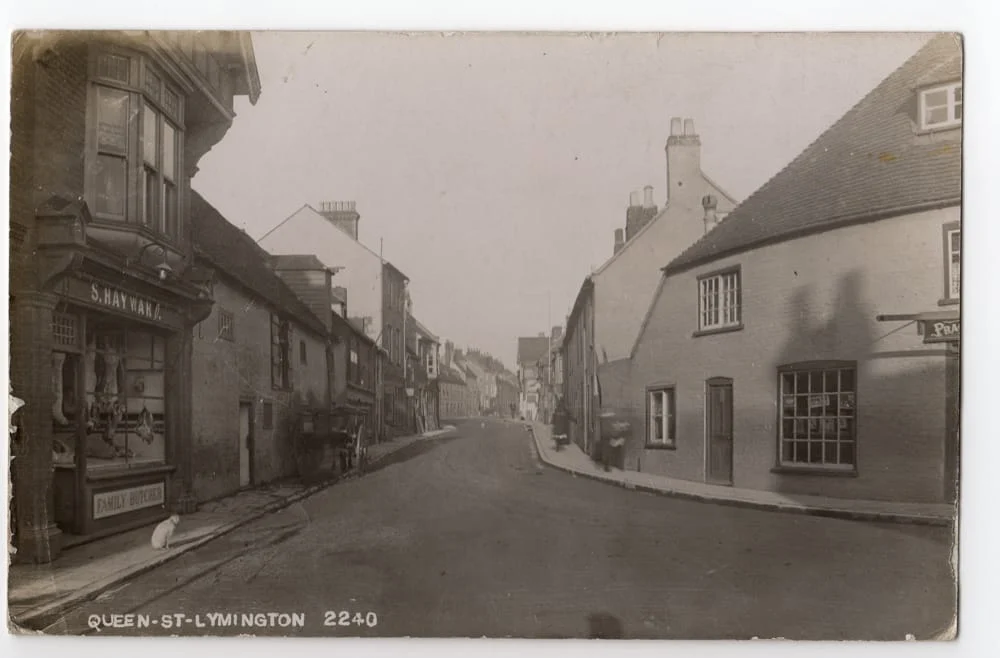 Black and white photo of a quiet street in Lymington, England, with old brick buildings, a shop sign reading S. Bayonar, and QUEEN ST-LYMINGTON 2240 written at the bottom. The street is empty.