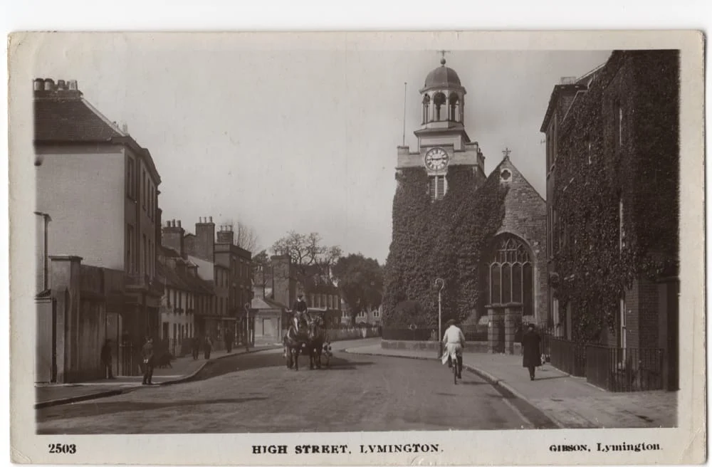Black and white vintage photo of High Street, Lymington, featuring a clock tower church, ivy-covered buildings, a horse-drawn carriage, and a person riding a bicycle along a quiet street.