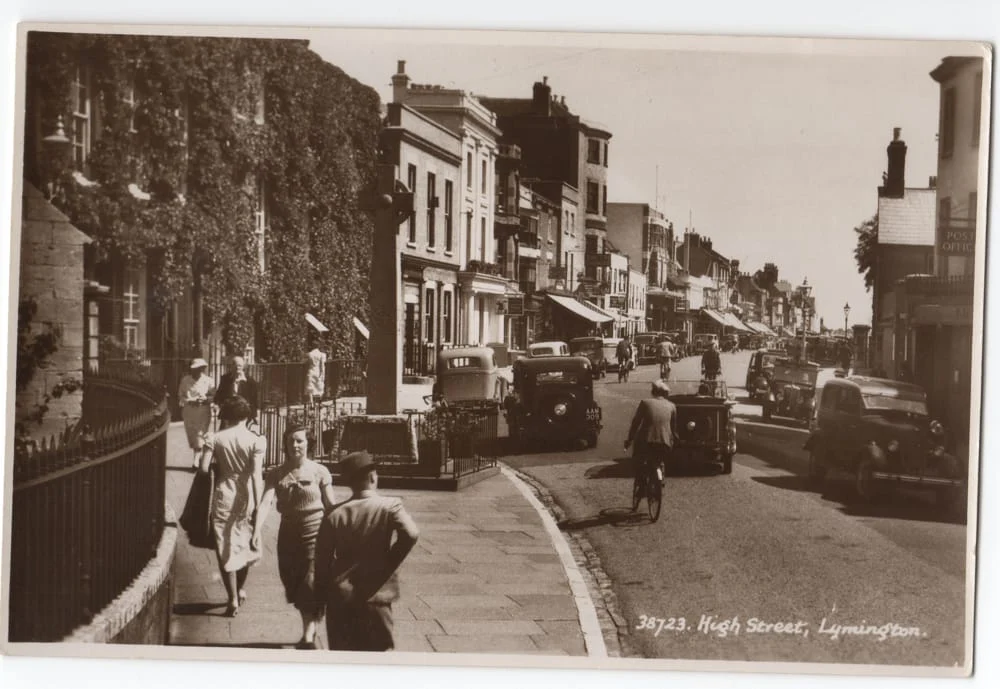 A black-and-white photo of High Street in Lymington, showing vintage cars, people walking on the pavement, and cyclists on the road, with old buildings and shops lining the street in sunlight.