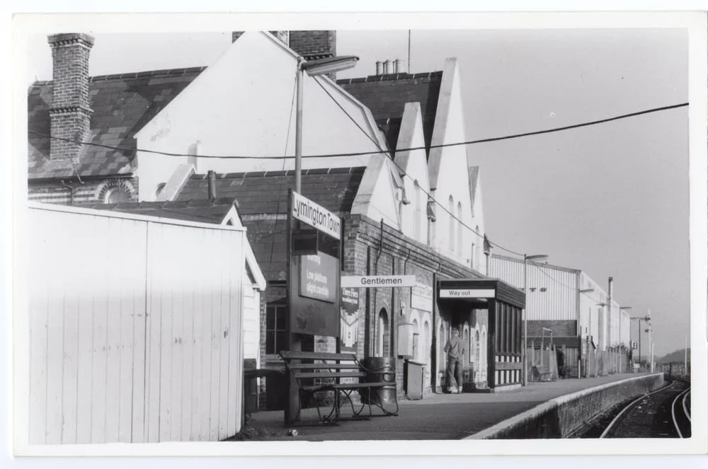 Black and white photo of a railway station platform with benches, a sign reading Gentlemen, and people waiting. Gabled buildings and a railway track run alongside the platform on a clear day.