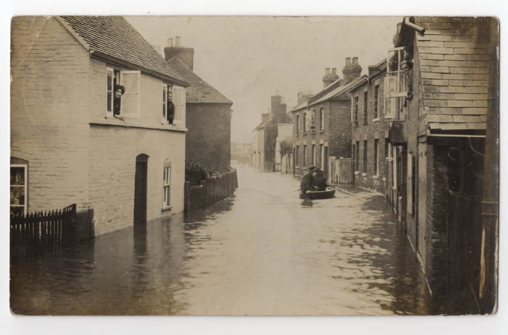 A sepia-toned photo of a flooded street with water reaching door level. A person rows a boat down the street, and a person looks out from an upstairs window of a brick house.