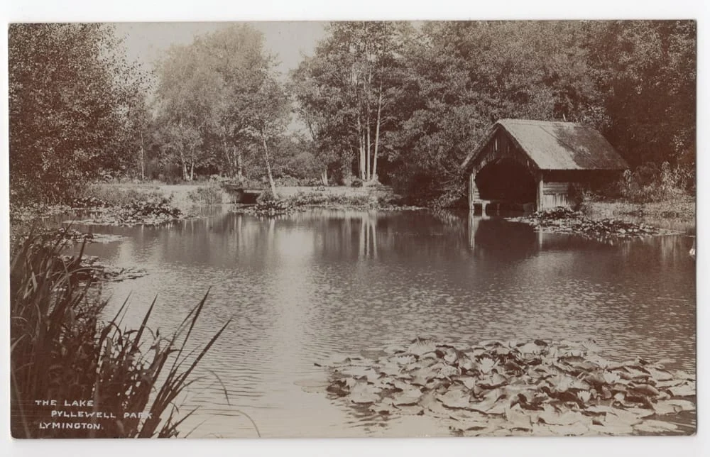 A calm lake surrounded by trees with water lilies in the foreground and a rustic wooden boathouse on the right. Text reads: “The Lake, PHILEWELL PARK, LYMINGTON.”.