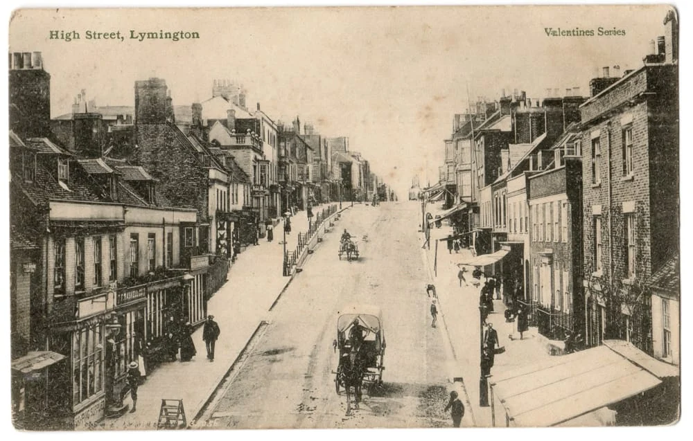 Sepia-toned vintage postcard of High Street, Lymington, showing a wide street lined with shops and buildings, horse-drawn carriages, and pedestrians. Text reads High Street, Lymington and Valentines Series.
