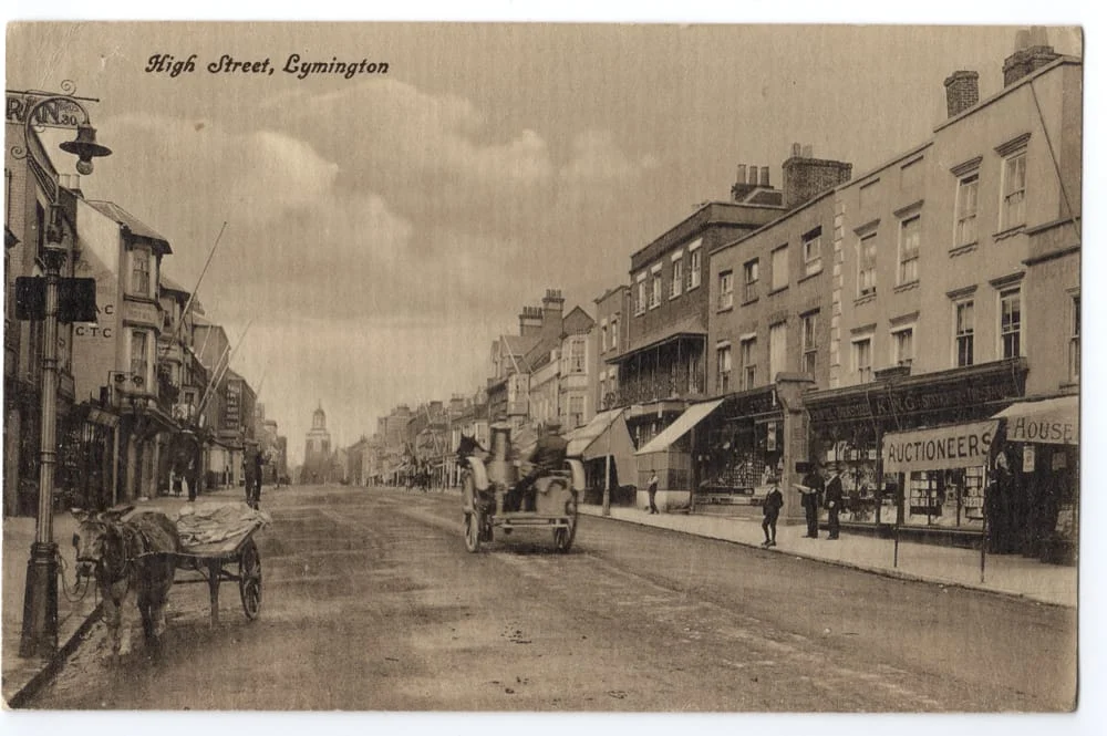 Sepia-toned photograph of High Street, Gymington, showing horse-drawn carts, pedestrians, and rows of shops and buildings lining both sides of a wide street under a cloudy sky.