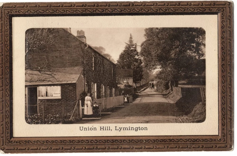 Sepia photograph of Union Hill, Lymington, showing a quiet road with brick houses on the left, a fence and trees on the right, and two women in aprons standing outside a house. The photograph is in a decorative frame.