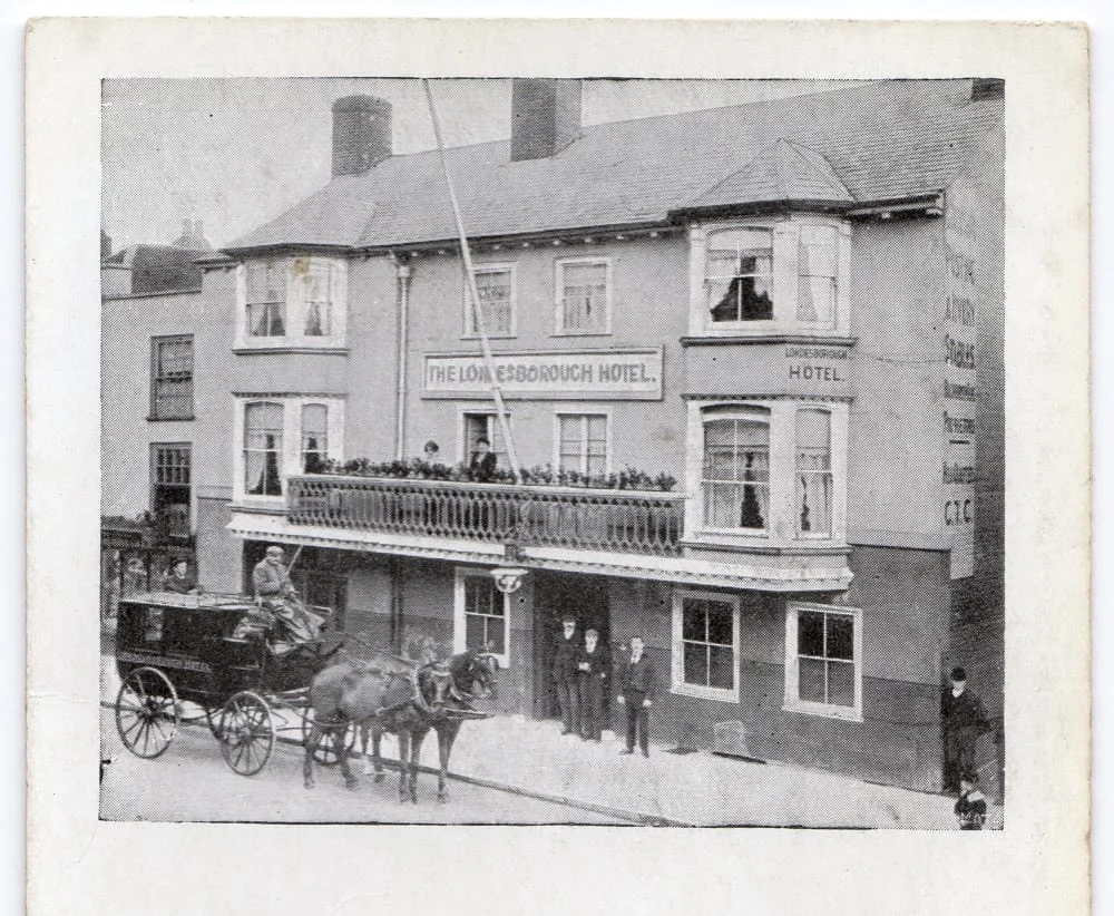 A black-and-white vintage photo shows The Loughborough Hotel with people standing on the balcony and outside the entrance. A horse-drawn carriage with a driver and passenger is parked in front of the building.