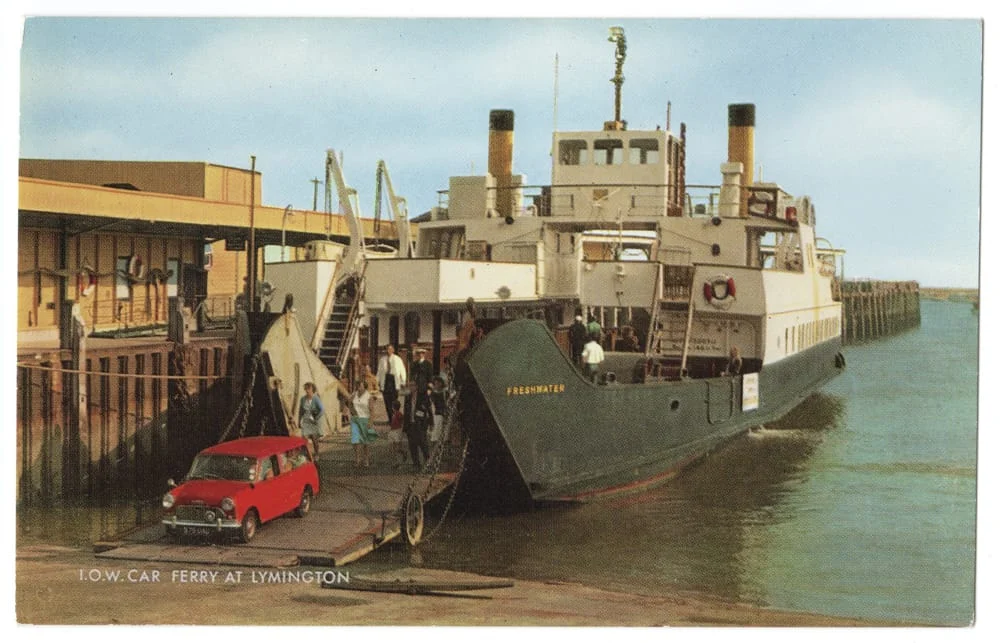 A vintage ferry named Freshwater docked at Lymington, with people and a red car boarding via a ramp; buildings and water are visible in the background.