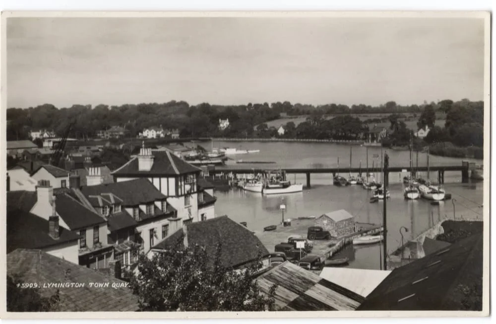 Black and white photo of Lymington Town Quay, showing a harbour with boats, riverside buildings, and a bridge, surrounded by trees and countryside in the background.