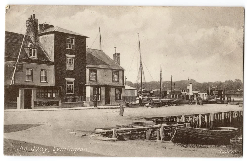 A sepia-toned vintage postcard shows The Quay in Lymington, with old buildings, boats moored along the waterfront, and a sign reading SHIPPING SUPPLIED. The scene has a peaceful, historic atmosphere.
