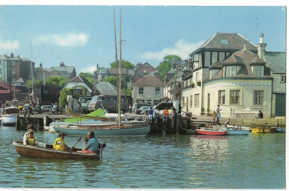 People in small boats float on a calm harbour, with sailing boats and colourful buildings in the background. Some people stand on the quay, and the sky is bright and clear.