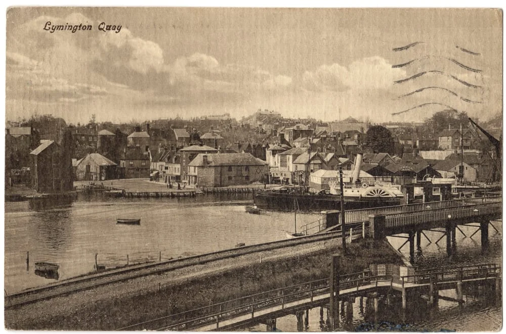 Sepia postcard of Lymington Quay showing historic buildings along the waterfront, a paddle steamer moored by the quay, small boats in the water, and wooden jetties in the foreground.