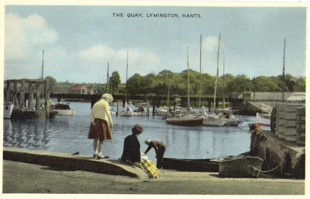 Two people sit near the water’s edge at a harbour with moored sailing boats, while another stands nearby. The scene is peaceful with trees and buildings in the background. Text reads: The Quay, Lymington, Hants.