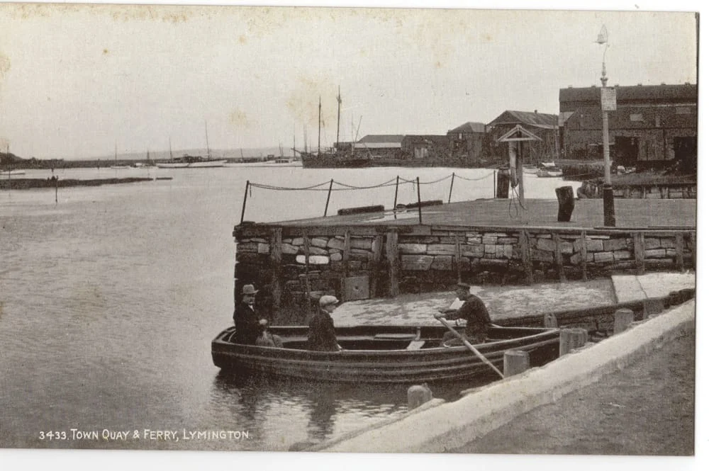 A vintage photograph shows people sitting in a small rowing boat near a wooden quay at Lymington. Several buildings, boats, and barrels are visible along the waterfront in the background.