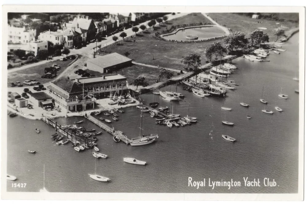 A black and white aerial photo of the Royal Lymington Yacht Club shows yachts and boats moored by the clubhouse, with nearby trees, parked cars, and waterfront buildings.