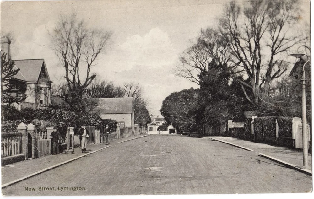 A black and white vintage photo of New Street in Lymington, showing a quiet, empty street lined with trees, houses, and fences on both sides, with a few people standing on the left pavement.
