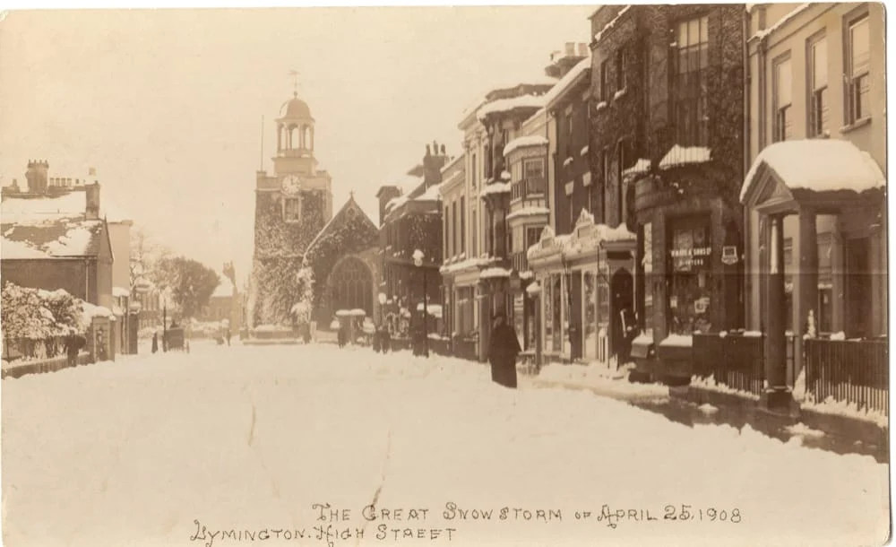 A sepia-toned photo of Lymington High Street covered in snow, with buildings lining both sides and a church with a clock tower in the background. Text at the bottom reads: “The Great Snowstorm of 25 April 1908.”.