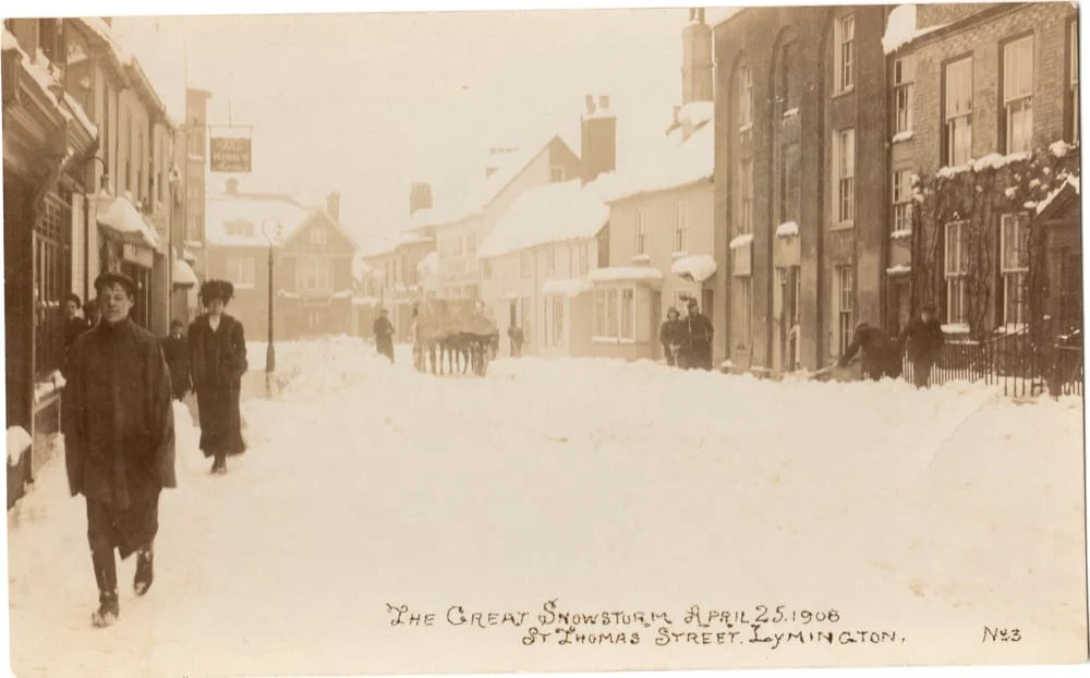 Sepia-toned photo of Lymington’s St Thomas Street after a heavy snowstorm on 25 April 1908. People walk on snow-covered streets, and tall buildings are lined with snow. Handwritten caption is visible at the bottom.