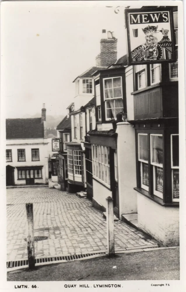 Black and white photo of Quay Hill in Lymington, showing old buildings along a cobbled street with a pub sign reading “MEW’S” featuring a drawing of a regal figure.