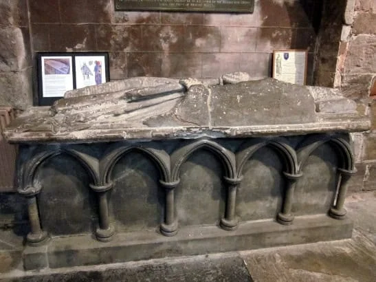 A weathered stone effigy of a medieval knight lies atop an ornate sarcophagus with arched detailing, set against a stone wall with framed historical information in the background.