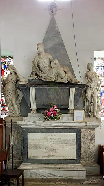 A marble monument features a reclining figure atop a tomb, flanked by two statues on each side, with stained glass windows in the background. Fresh flowers and a framed plaque are placed on the tomb.