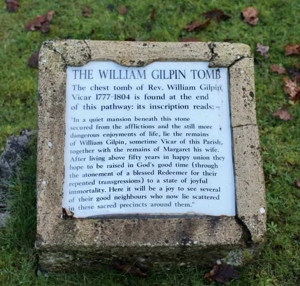 A weathered stone plaque on grass marks The William Gilpin Tomb. The inscription honours Rev. William Gilpin, vicar 1777-1804, describing his peaceful rest and hope for resurrection with loved ones.