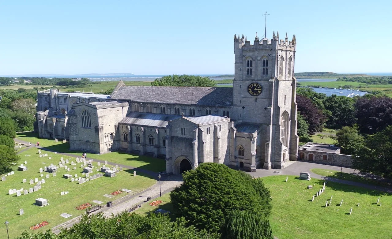 A large stone church with a clock tower stands in the centre of a green churchyard with gravestones, surrounded by trees under a clear blue sky. A river and fields are visible in the background.