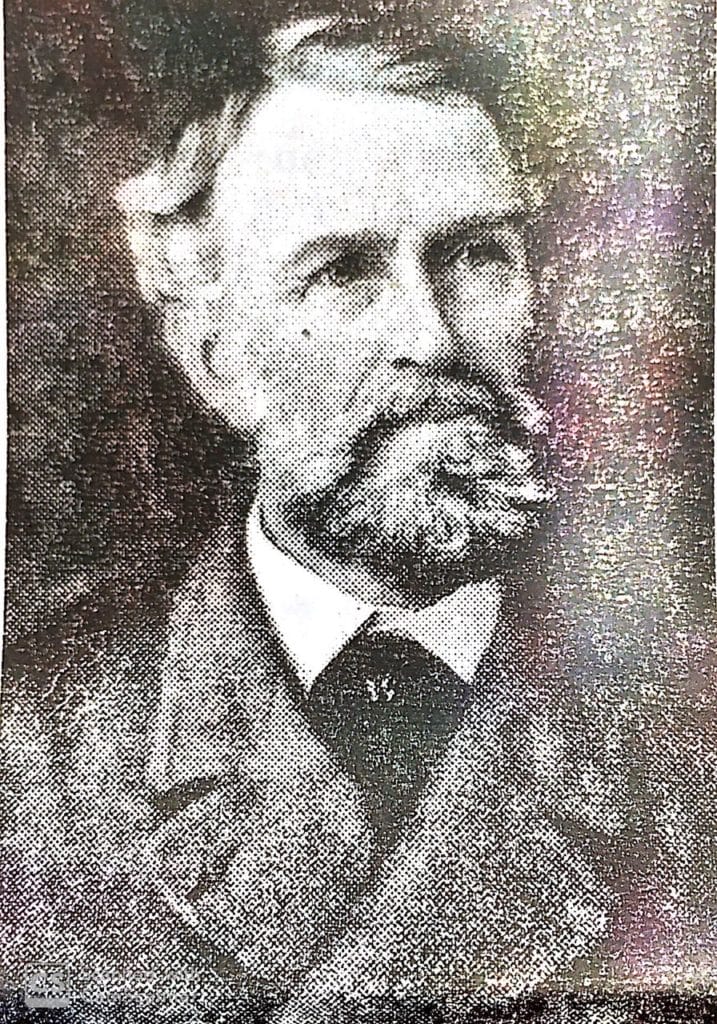 Black-and-white, grainy portrait of an older man with a beard and moustache, wearing a suit and tie, looking slightly to the side. The image appears to be historic or from an old photograph.