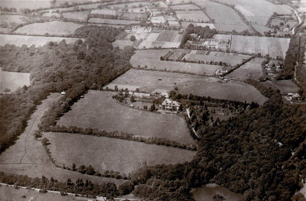 A black-and-white aerial photo shows a rural landscape with patchwork fields, wooded areas, and a large house surrounded by trees, roads, and farmland.