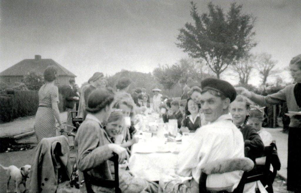 A black and white photo showing a group of people, including children and adults, sitting and standing around a long outdoor table having a meal. Trees and houses are visible in the background under a cloudy sky.