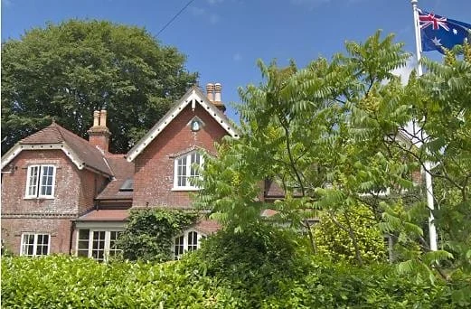 A red-brick house with white-trimmed windows and a steep roof stands behind green bushes and trees. An Australian flag is visible on a pole to the right, and the sky above is clear and blue.