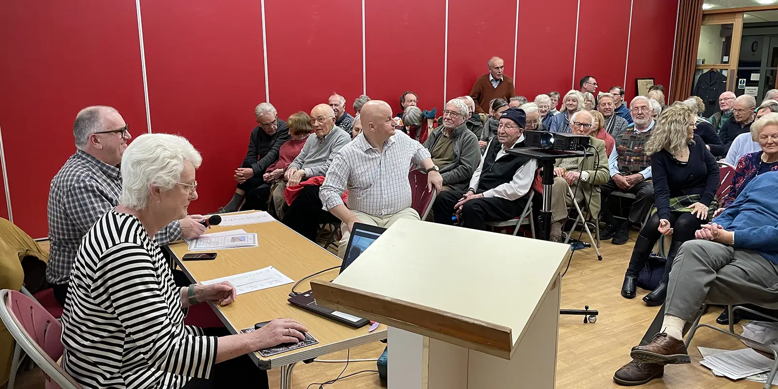 A group of older adults sits closely together in a brightly lit room with red walls, facing a table where two people are seated with papers and a laptop. A white lectern is visible in the foreground.